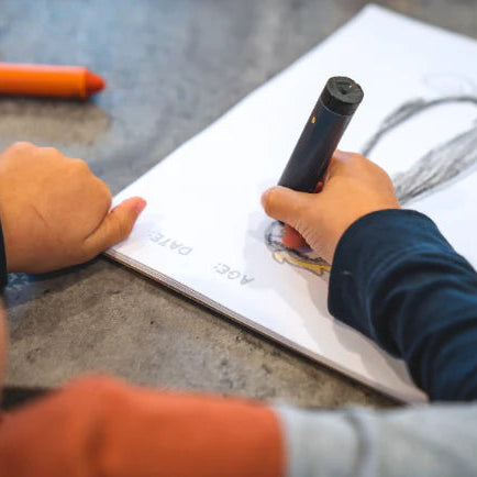 Child holding a black marker over a notebook with drawings on a gray surface